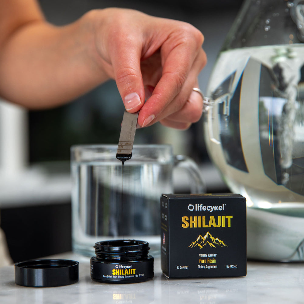 A hand holding metal dosage measurement with Lifecykel Shilajit liquid syrup in a jar and packaging box and lid on the side