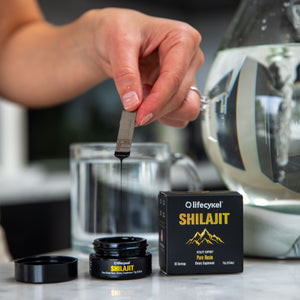 A hand holding metal dosage measurement with Lifecykel Shilajit liquid syrup in a jar and packaging box and lid on the side