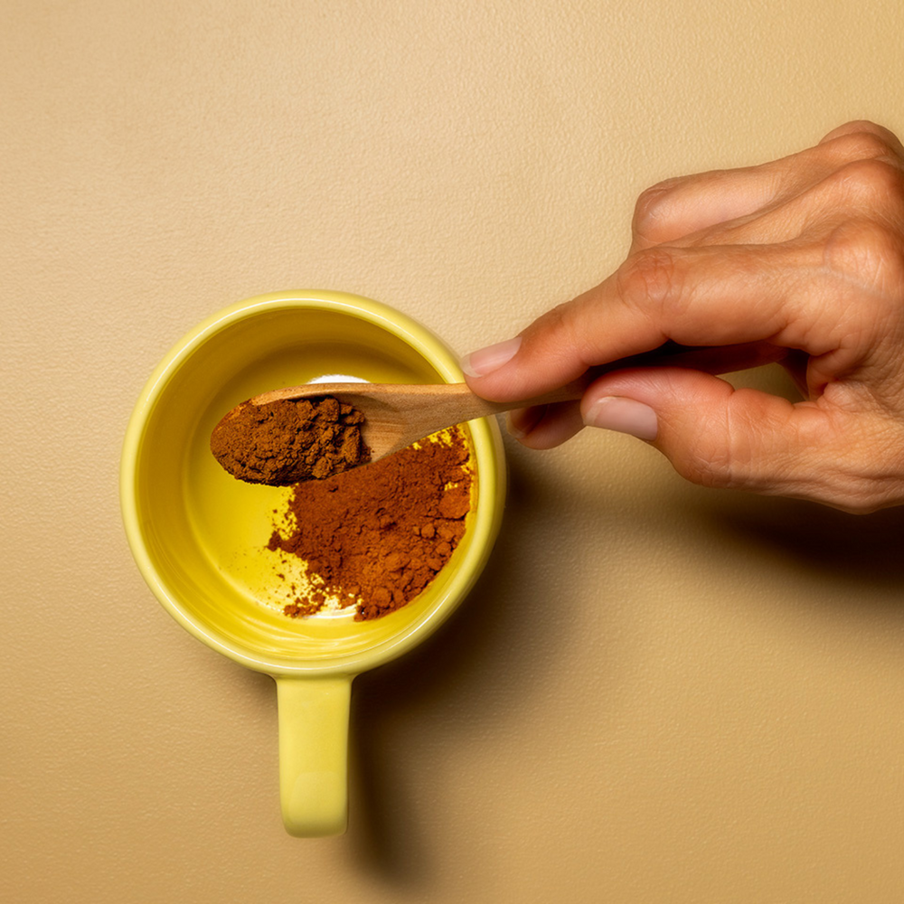 A hand with wooden spoon pouring the coffee powder on the yellow ceramic mug in plain yellow background
