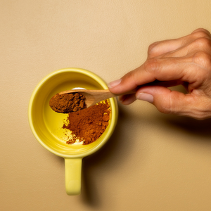 A hand with wooden spoon pouring the coffee powder on the yellow ceramic mug in plain yellow background
