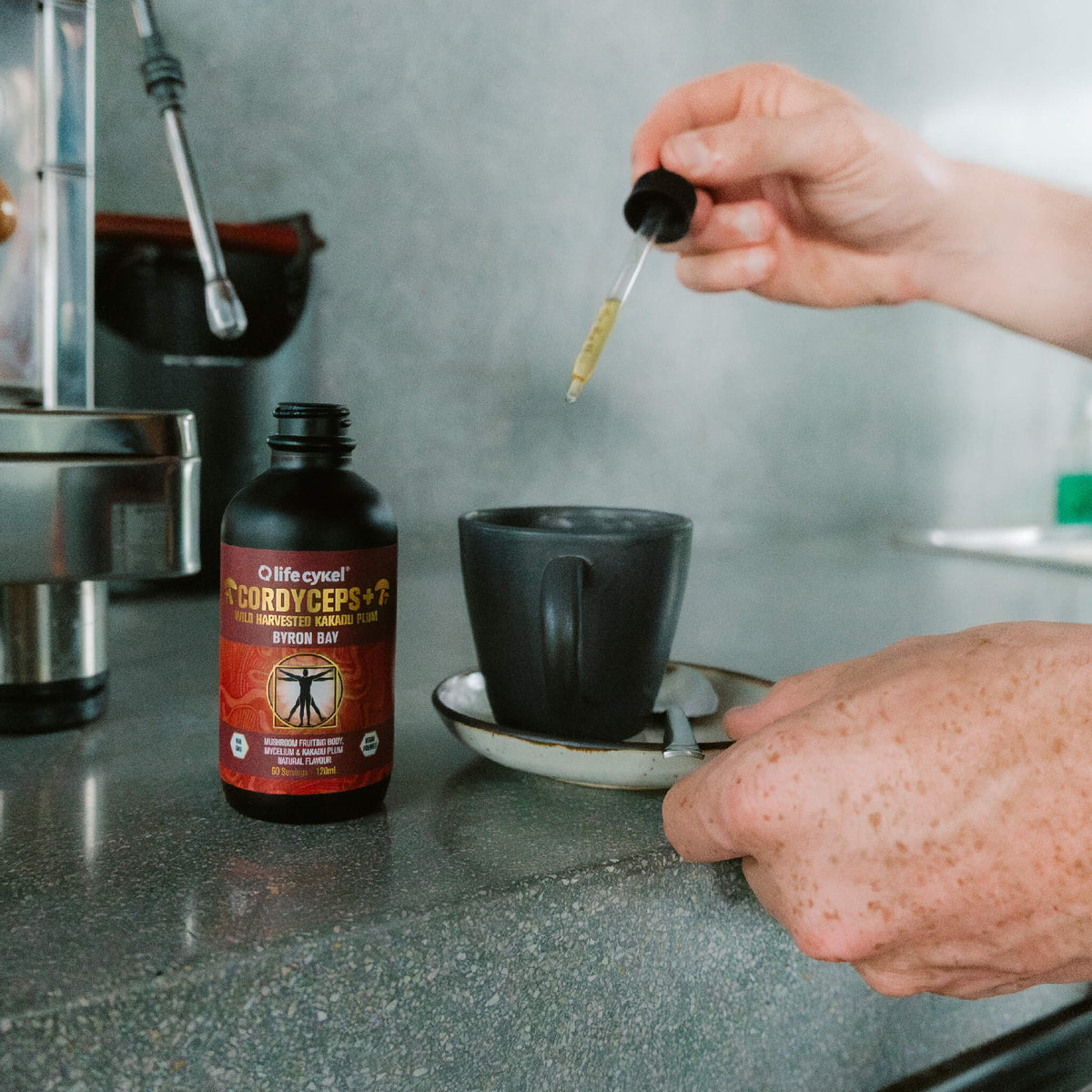 A Lifecykel Cordyceps mushroom extracts supplements with a hand with dropper pouring liquid to black ceramic mug 